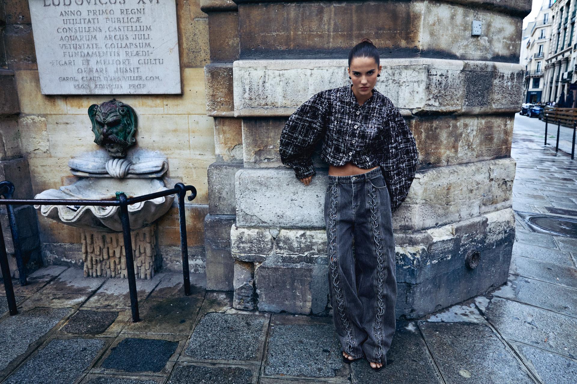 A woman stands in front of a stone wall in a city wearing an outfit from the Danish brand ROTATE. She is wearing a cropped, monochrome tweed style jacked with voluminous sleeves and a pair of grey jeans with lace up detailing down the front.